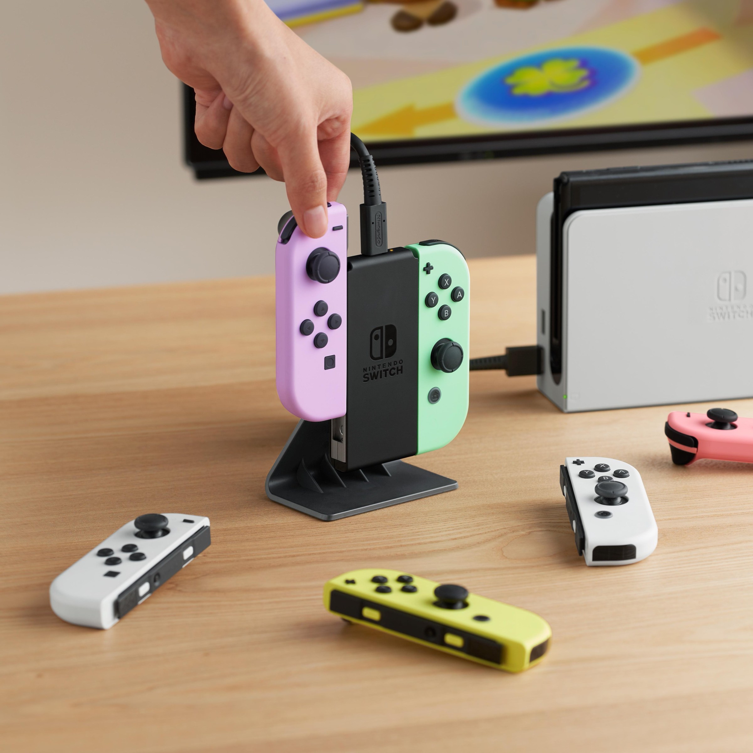 A photo of a Joy-Con Charging Stand for Nintendo Switch. A hand slides a Joy-Con into one side of the stand, which is plugged into a Switch dock. Other Joy-Cons are scattered around it.